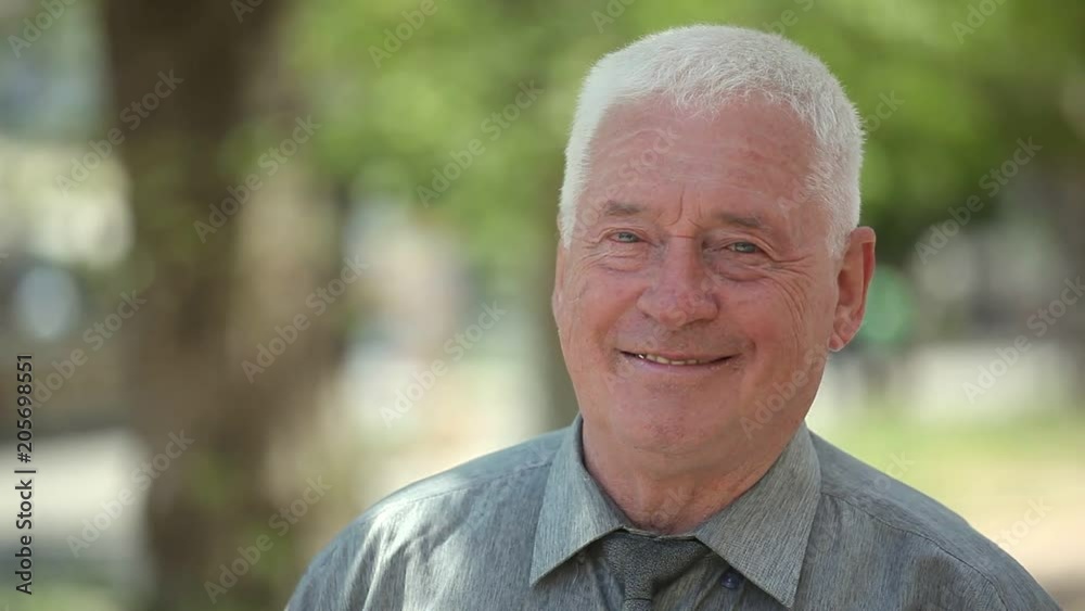 Portrait of a cheery senior man with a short haircut in a blue shirt with a wrinkled face. He stands and smiles sincerely in a street in summer. Portrait of a cheery senior man with a short haircut in a blue shirt with a wrinkled face. He stands and smiles sincerely in a street in summer.