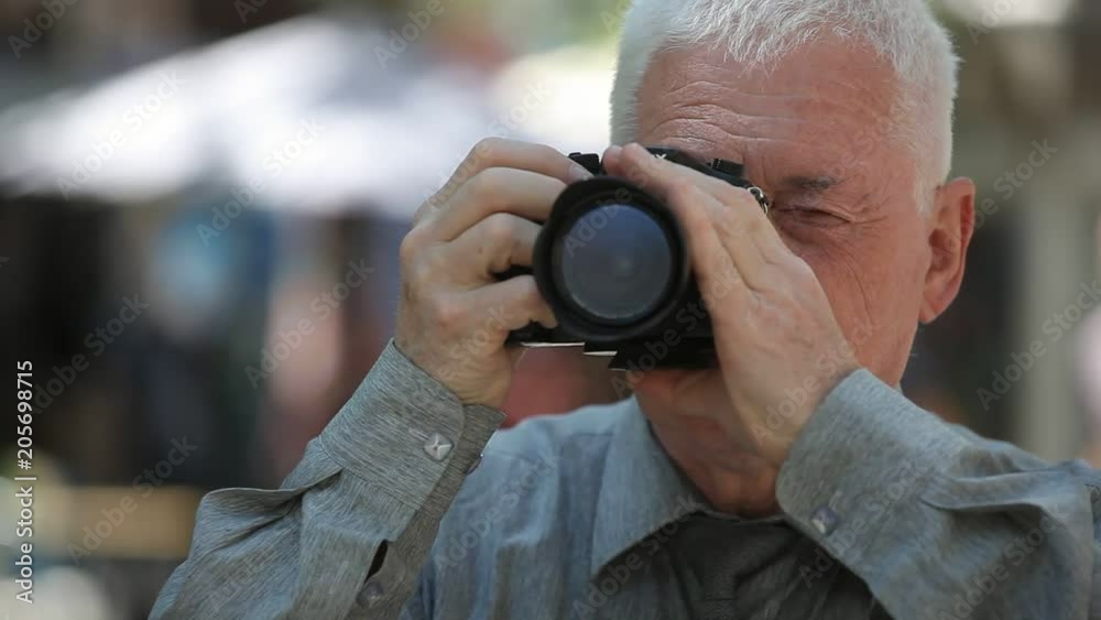 Bokeh shot of a grey-headed man with a crew haircut in a grey shirt standing in a street, raising his camera and taking photos in summer 