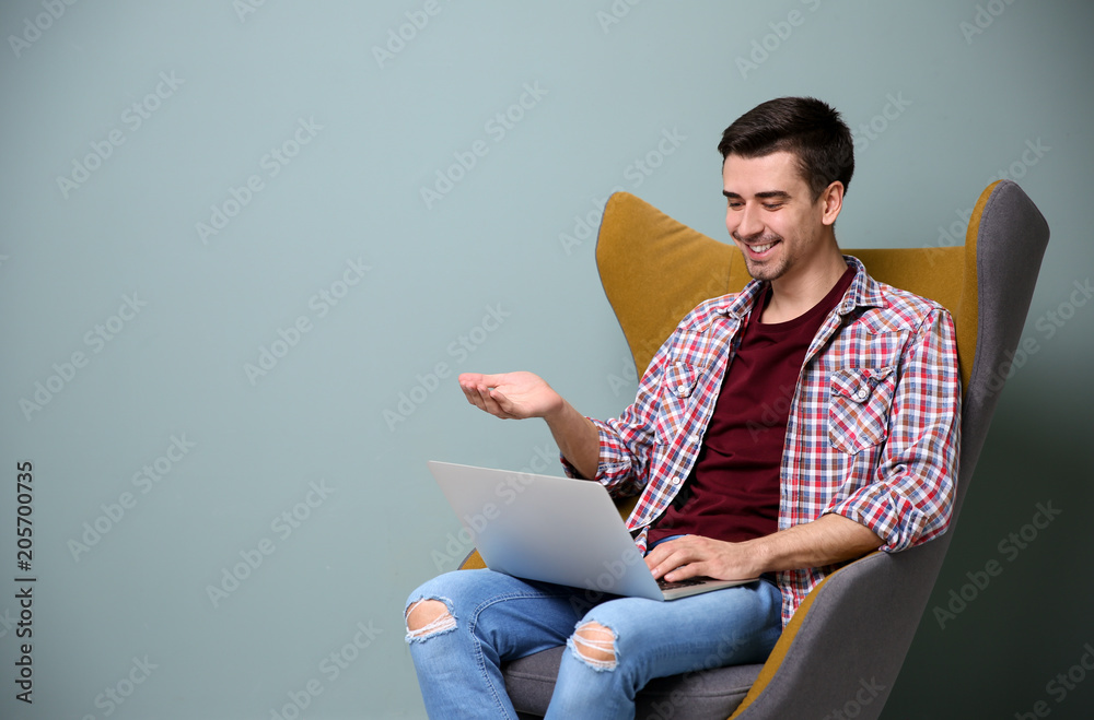 Young blogger with laptop sitting in armchair against color wall