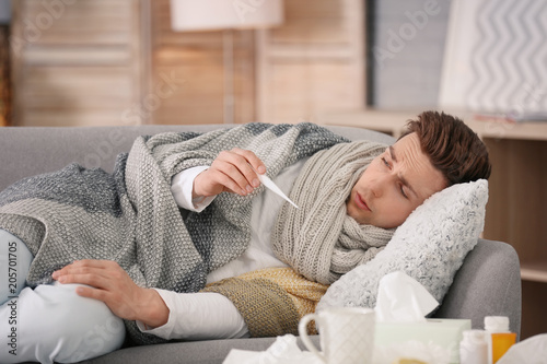 Canvas Print Sick young man with thermometer suffering from cold on sofa at home