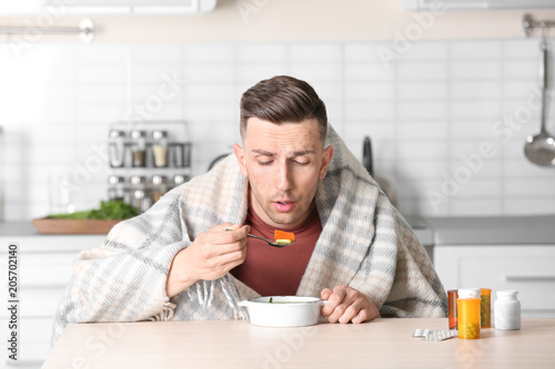 Sick young man eating broth to cure cold at table in kitchen