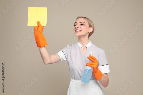 Young chambermaid with rag and detergent on color background