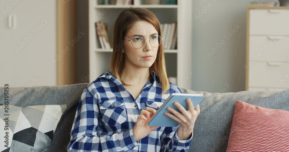 Young woman in glasses and motley shirt scrolling and taping on the tablet computer on the sofa at home, then thinking. Inside Young woman in glasses and motley shirt scrolling and taping on the tablet computer on the sofa at home, then thinking. Inside