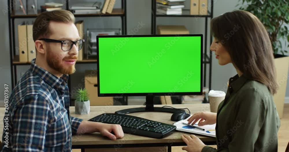 Back view on the couple of co-workers sitting in the office at the table and working in front of the computer green screen, then turning to the camera and showing ok gesture. Chroma key. Indoor