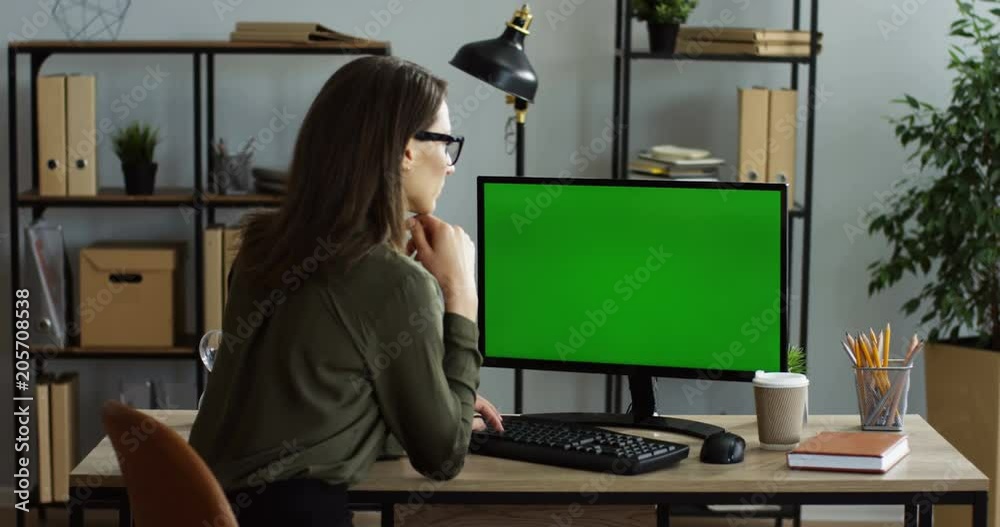 Rear view on the caucasian woman sitting at the office desk and taping on the keyboard of the computer with green screen, thinking while texting. Chroma key. Indoors