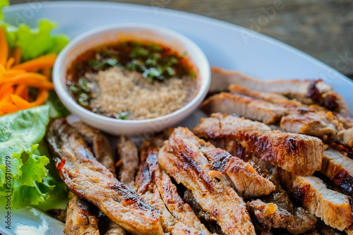 Grilled pork in a white dish with vegetables and dipping sauce on a wooden table.
