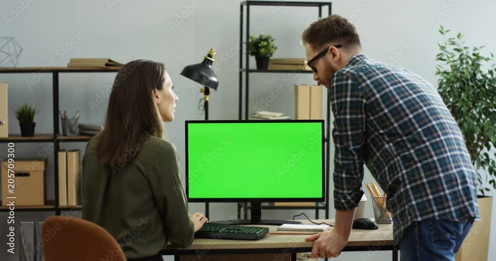 View from behind on the woman sitting at the desk and working in front of the computer green screen, man standing next to her and they talking. Chroma key. Inside