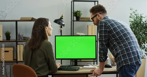 View from behind on the woman sitting at the desk and working in front of the computer green screen, man standing next to her and they talking. Chroma key. Inside