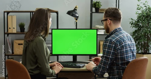 Rear view on the male and female office workers sitting at the desk and working in front of the computer green screen, discusing their project. Chroma key. Indoor