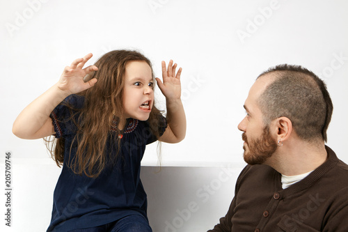 Studio shot of funny emotional little girl with messy hairstyle grimacing and making fearful gesture with both hands, trying to scare her attractive bearded father while playing together indoors