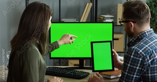 View from behind on the office workers sitting at the table in front of the computer green screen, man holding a tablet device and they watching something on it. Chroma key. Indoors