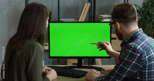 View from the back on the male and female office workers sitting at the desk in front of the computer and they watching something on its green screen. Chroma key. Inside