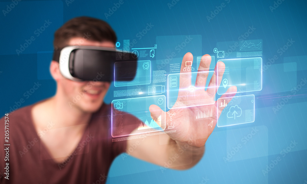Young impressed man wearing virtual reality goggles with blue squares containing data at his fingers Young impressed man wearing virtual reality goggles with blue squares containing data at his fingers