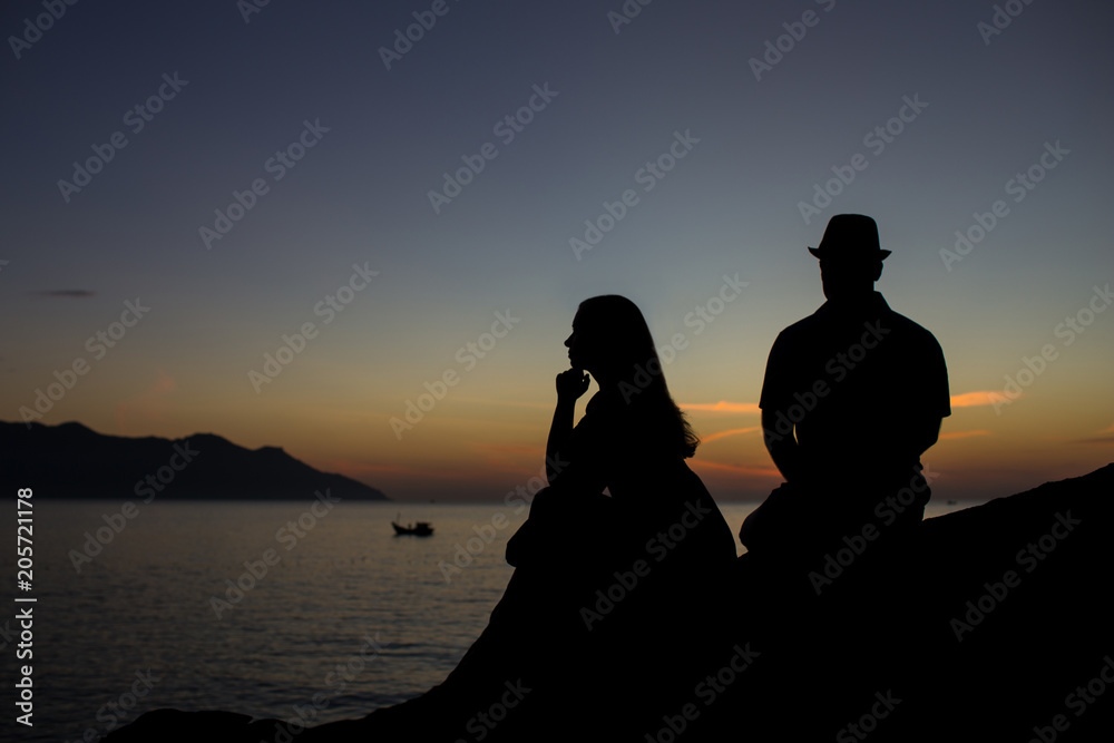 Silhouette of young couple are sitting on a rock at sunrise near the sea.