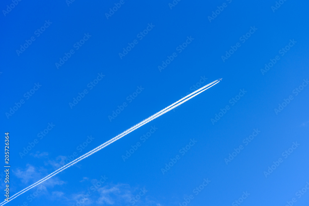 A distant aircraft with long diagonal condensation trails or contrails on a blue sky day A distant aircraft with long diagonal condensation trails or contrails on a blue sky day