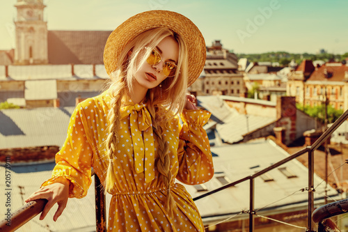 Outdoor portrait of young beautiful girl wearing trendy yellow color sunglasses, straw boater hat, polka dot dress posing in street of european city. Summer fashion concept. Copy. empty space 