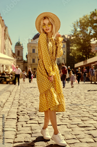 Outdoor full body portrait of young beautiful fashionable girl wearing trendy yellow color sunglasses, straw boater hat, polka dot dress posing in street of european city. Summer fashion concept