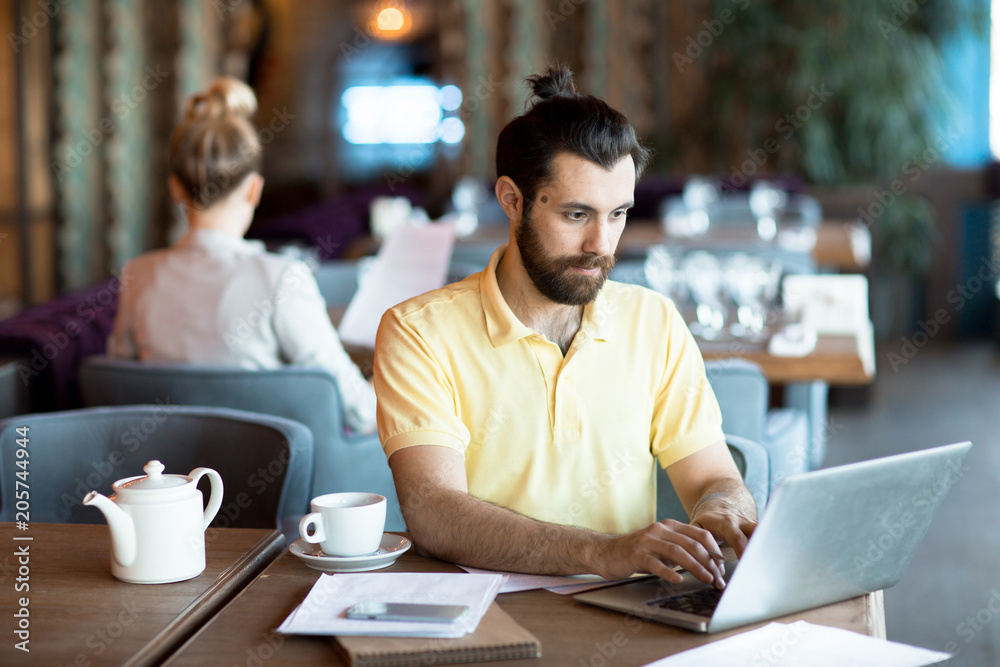 Busy young man in yellow shirt sitting by table in front of laptop, networking or preparing for business seminar