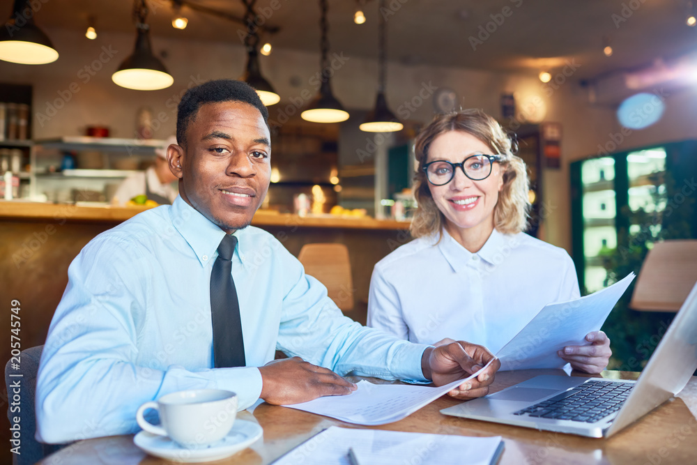 Two confident financiers looking at camera while sitting by table in cafe and working with papers