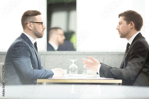 Side view of two young businessmen sitting by table in front of one another and discussing new business