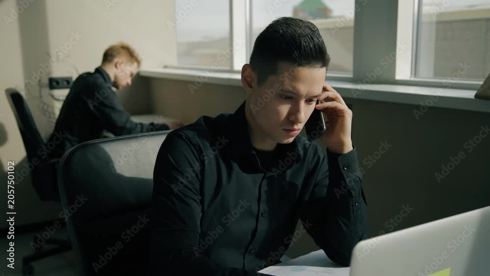 Male office worker talking on phone and his colleague is sitting at the background. Concept of: business people, finanacial planning, office interior, digital technologies, business communication Male office worker talking on phone and his colleague is sitting at the background. Concept of: business people, finanacial planning, office interior, digital technologies, business communication