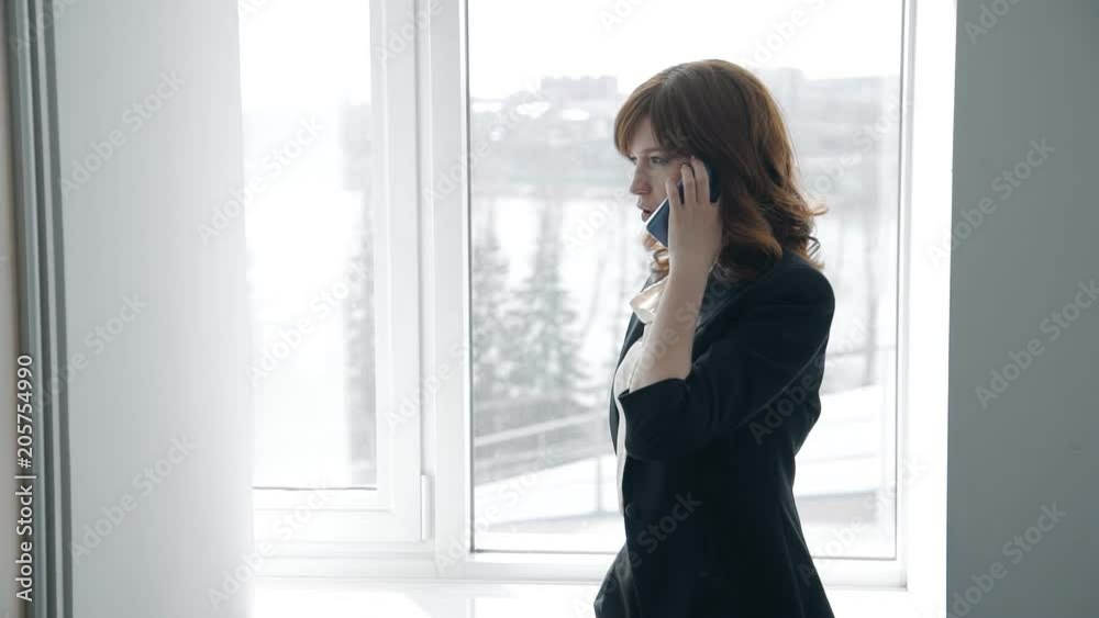 Attractive businesswoman is talking on phone by window in modern office, successful person making call, walking, discussing business in company. Concept: business conversation, professional