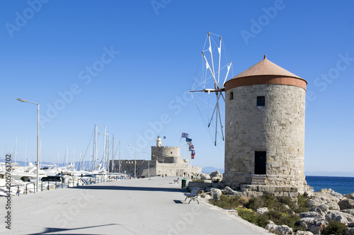 Ancient windmills on stony Rhodes coastline in harbor, old historic buildings, place of interest, blue sky