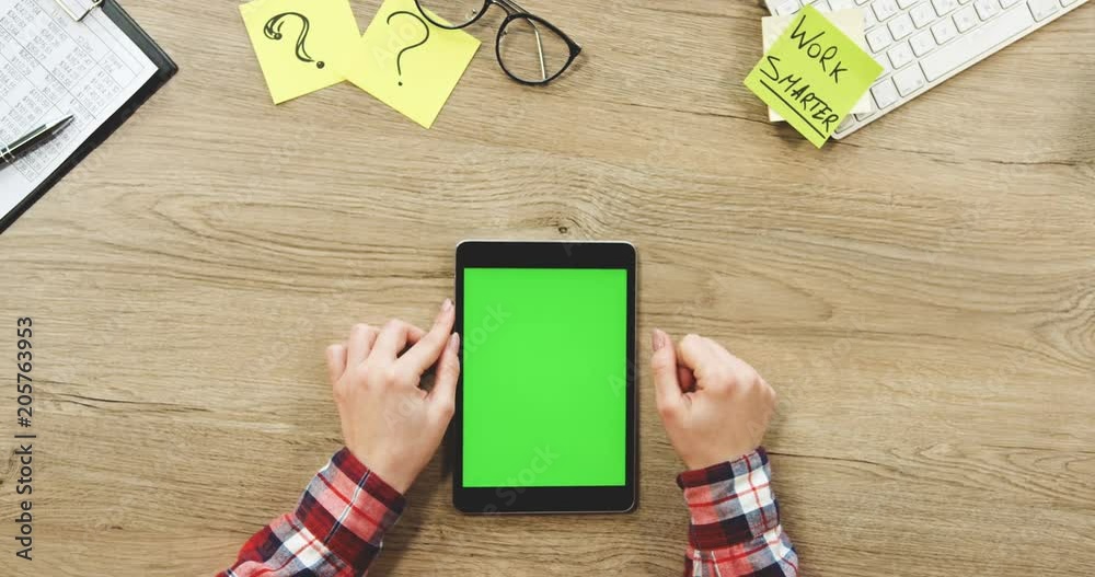 Close up of the female hands taping on the black vertical tablet device with a green screen on the wooden office table. Work smarter note. Chroma key. Top view.
