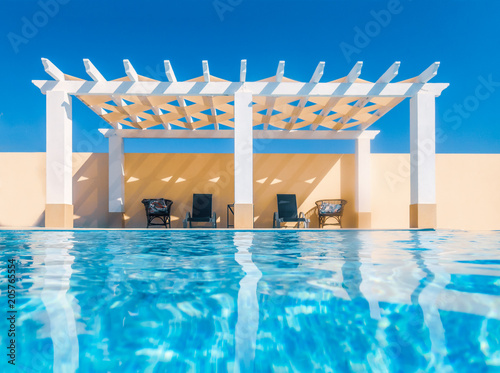 White poolside pergola, gazebo taken from the swimming pool. The reflection of the pergola can be seen in the water