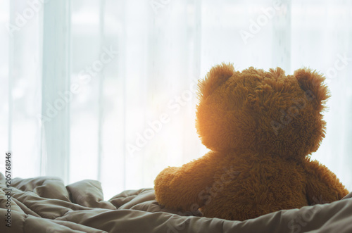 brown teddy bear sitting on bed looking on windows with sunlight in the bedroom on morning.