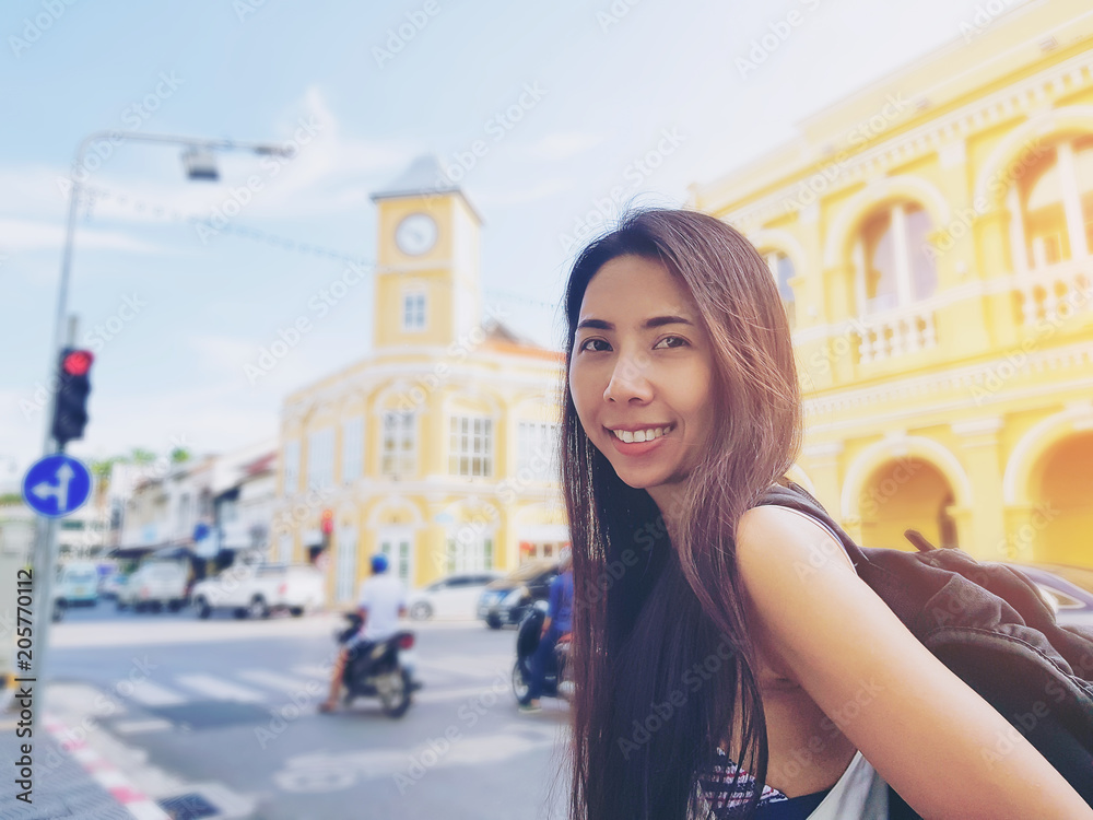 Young Asian woman traveler with a backpack on her shoulder out sightseeing or enjoying the view from in Phuket Old Town. Thailand.  Hipster girl smile while looking camera. Travel Holiday Asia Concept
