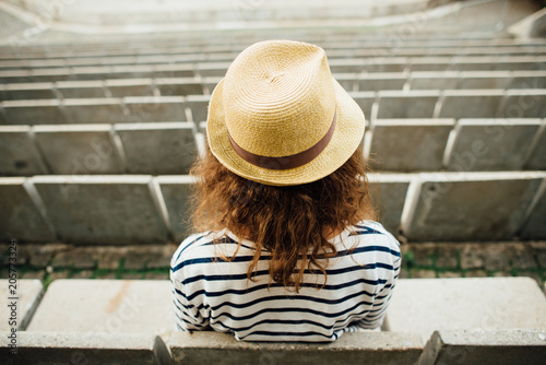 Young woman in a hat sitting in the amphitheatre in the park alone, photographed from behind