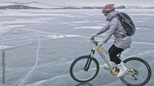 Woman is riding bicycle on the ice. The girl is dressed in a silvery down jacket, cycling backpack and helmet. Ice of the frozen Lake Baikal. The tires on the bicycle are covered with special spikes photo