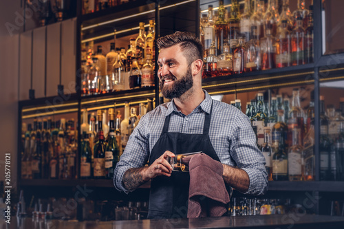 Cheerful stylish brutal barman is cleaning the glass with a cloth at bar counter background. photo