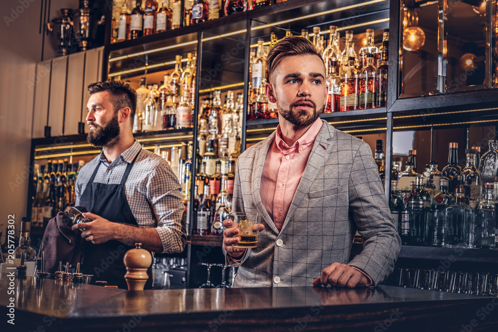 Stylish handsome male in an elegant suit holds a glass of whiskey at bar counter background.