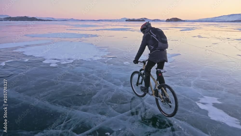 Man is riding a bicycle on ice. The cyclist is dressed in a gray down jacket, backpack and helmet. Ice of the frozen Lake Baikal. The tires on the bicycle are covered with special spikes. The traveler Man is riding a bicycle on ice. The cyclist is dressed in a gray down jacket, backpack and helmet. Ice of the frozen Lake Baikal. The tires on the bicycle are covered with special spikes. The traveler