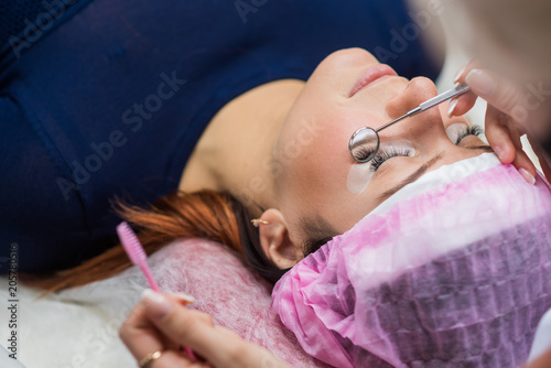 Having eye lash extension procedure. Young woman lying on the back in a salon, stilist holding round mirror showing eyelashes extension in the mirror.