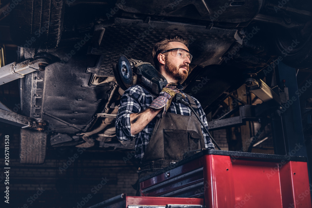 Mechanic in a uniform and safety glasses holds an angle grinder while standing under lifting car in a repair garage. 