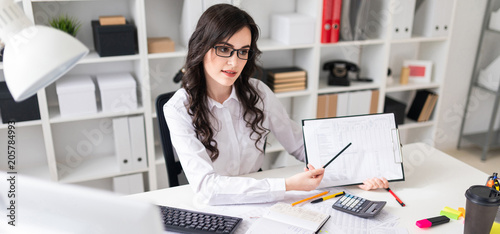 A beautiful young girl is sitting at a table in the office and pointing a pencil at the information in the document.