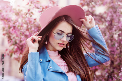 Young beautiful fashionable girl wearing stylish blue color aviator sunglasses, pink suede hat, earrings, biker jacket. Model posing in street with flowering trees. Spring fashion concept. 
