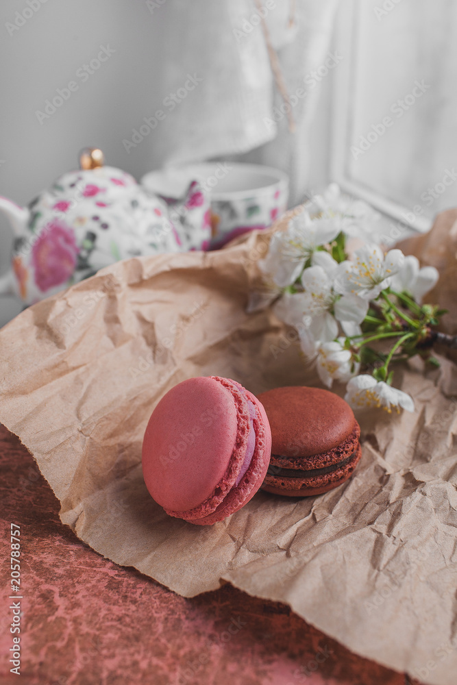 French Colorful pink and brown Macarons Pastel on old craft paper on a pink and brown Background with with a blooming cherry branch, tea pot and cup in pink colors style Provence near white window French Colorful pink and brown Macarons Pastel on old craft paper on a pink and brown Background with with a blooming cherry branch, tea pot and cup in pink colors style Provence near white window
