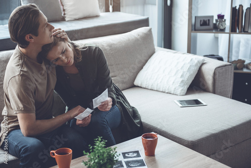 Frustrated young woman is suffering after miscarriage. She is looking at ultrasound pictures of her unborn baby and crying. Man is sitting and embracing her with love. Copy space  photo