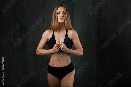 Image of muscular young female athlete wearing black sport wear standing looking in camera on dark background. Woman bodybuilder resting after workout.