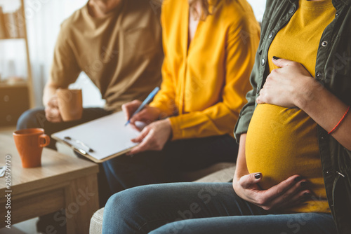 Focus on pregnant woman touching her abdomen with gentleness. Married couple is putting signature on document while sitting on background. Surrogate motherhood concept  photo