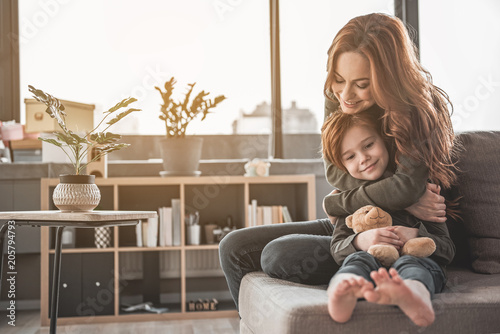 Full length portrait of affectionate mom cuddling her kid with delight. Small girl is holding her teddy bear with her legs stretched. Copy space in left side photo