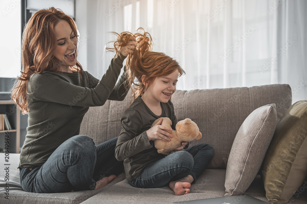 Full length portrait of laughing mom and kid fooling around on their sofa. Mother playfully holding her child red curls. Happy little girl is hugging teddy bear with enthusiasm