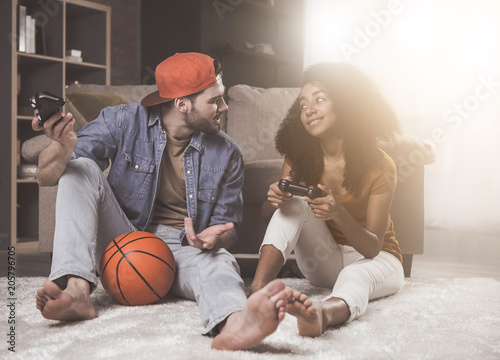 Fun together. Full length of pleasant stylish romantic guy and girl are talking and playing on console. They are sitting on floor at home with basket ball