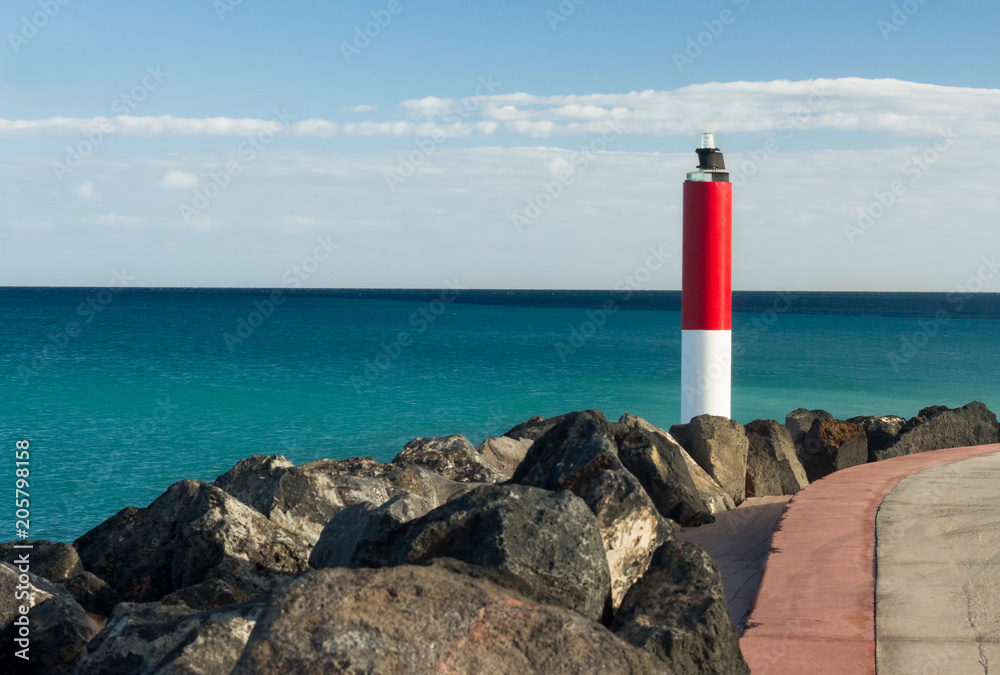 A bright red white lighthouse, surrounded by large stones, on the background of the blue Atlantic ocean and the breeze on the water