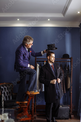 Bilde på lerret Mature professional tailor putting himself to a high stool in order to present an elegant look of bespoken coat to young male client in exclusive atelier studio