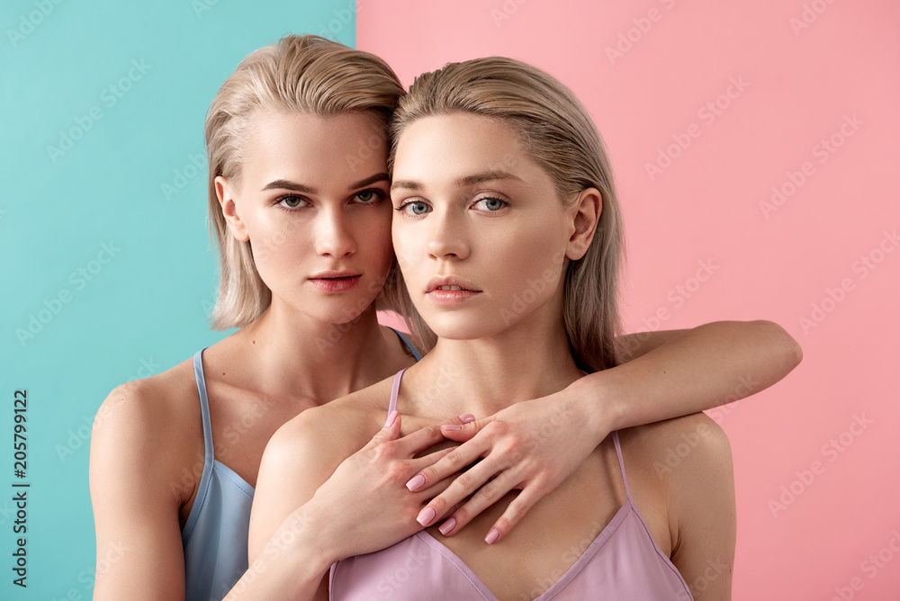 Friendship concept. Portrait of blonde women staring at camera with tranquility while cuddling. Isolated on blue and pink background Friendship concept. Portrait of blonde women staring at camera with tranquility while cuddling. Isolated on blue and pink background